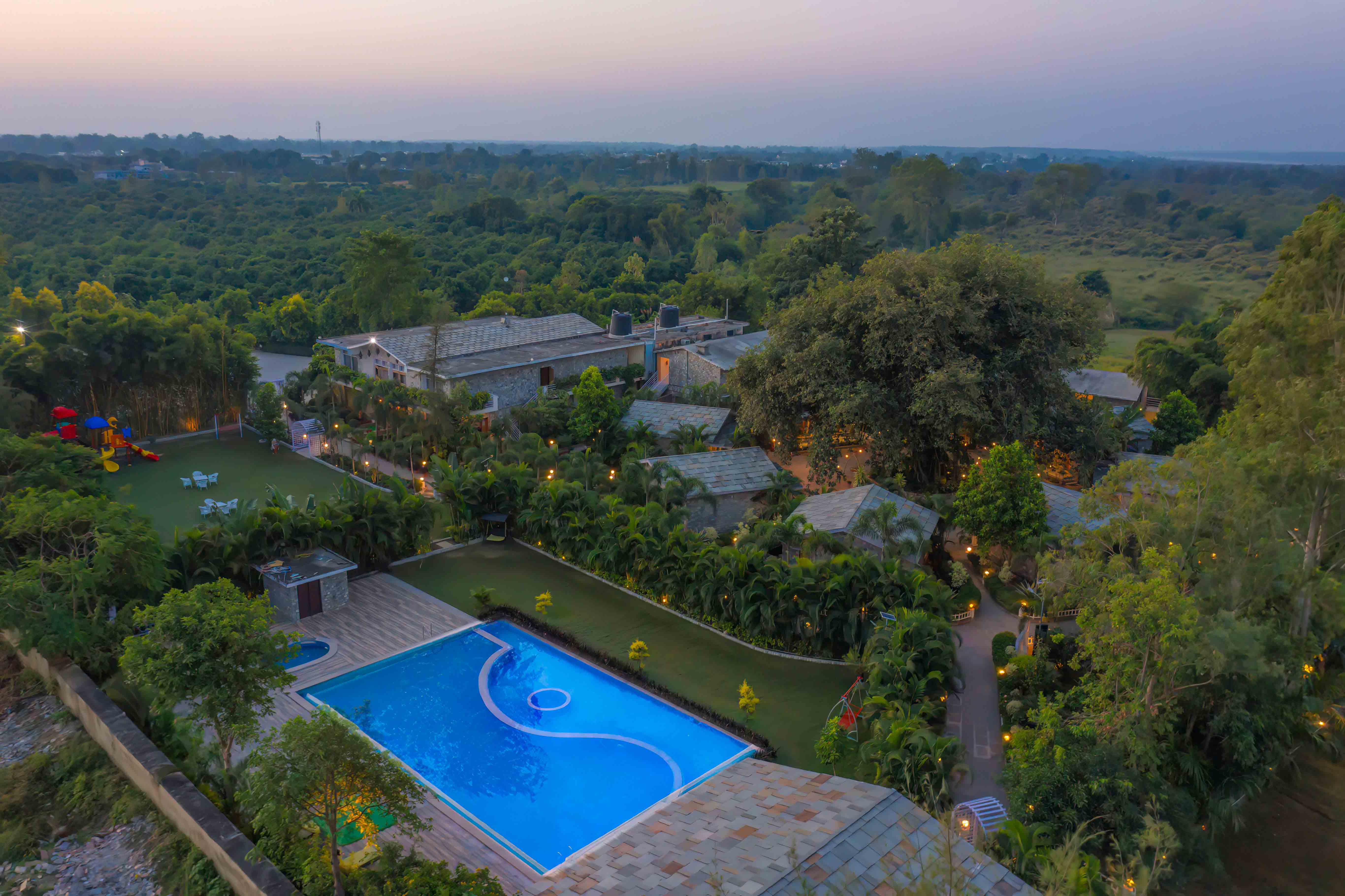 Aerial evening view of The Banyan Retreat with its glowing swimming pool amidst dense Corbett forest